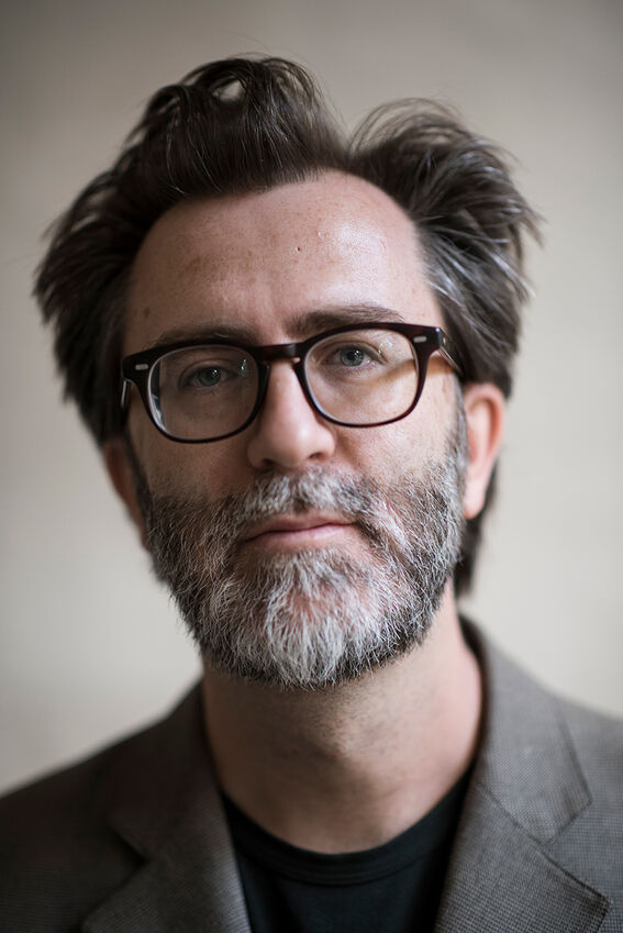 Portrait of a middle‑aged man with glasses and greying beard, wearing a brown jacket against a plain background