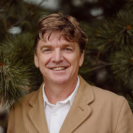 Smiling middle‑aged man in tan blazer and white shirt standing among evergreen branches outdoors