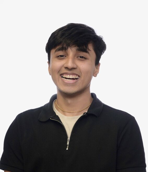 Young man smiling at the camera, wearing a black top and gold necklace on a plain light background