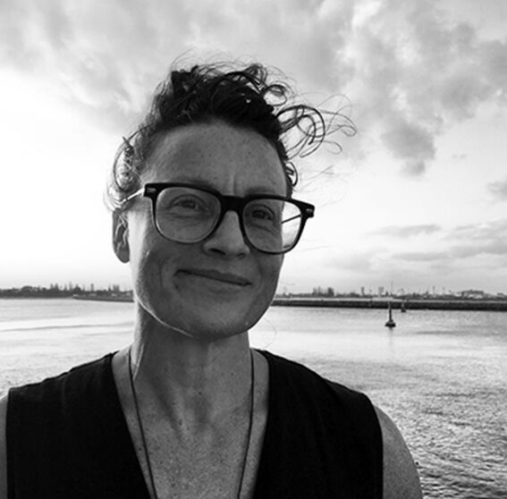 Black and white portrait of a smiling person with glasses by calm water under cloudy sky
