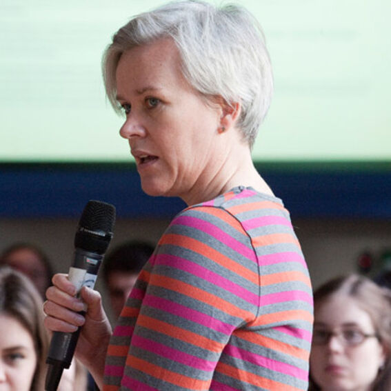 Short-haired adult with grey hair holding a microphone, speaking to an audience, wearing a striped pink and grey top.