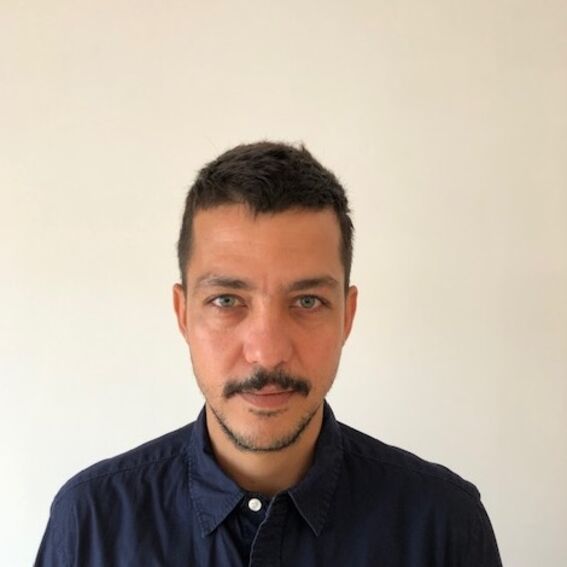 Man with short dark hair and moustache in dark blue shirt against plain light wall