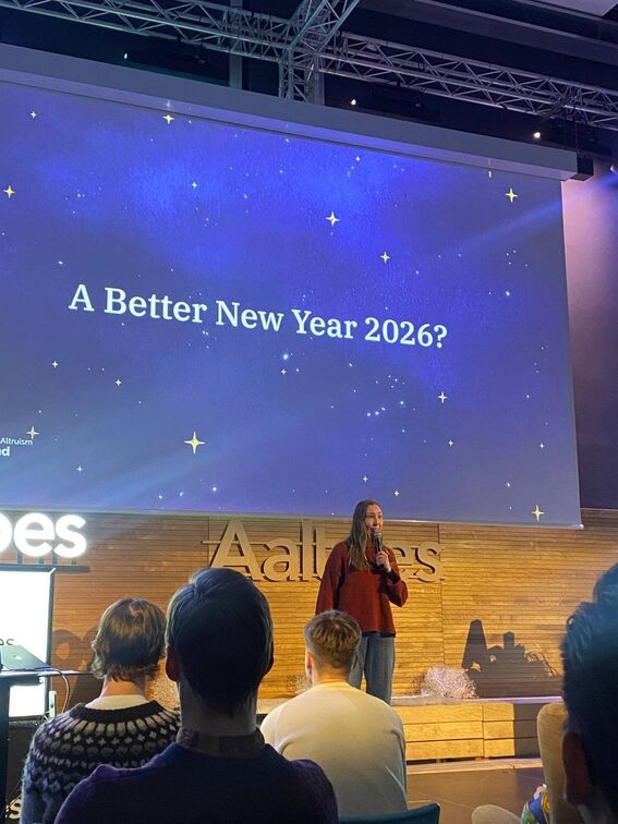 Speaker on stage with mic under slide reading “A Better New Year 2026?” in starry blue theme, audience seated