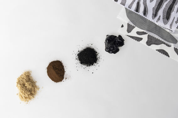 Four piles of natural pigments from beige to black beside grey patterned fabric samples on white surface