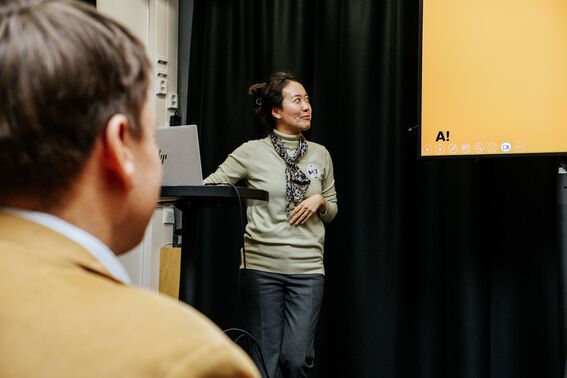 Woman in green jumper presents beside yellow screen to seated audience member