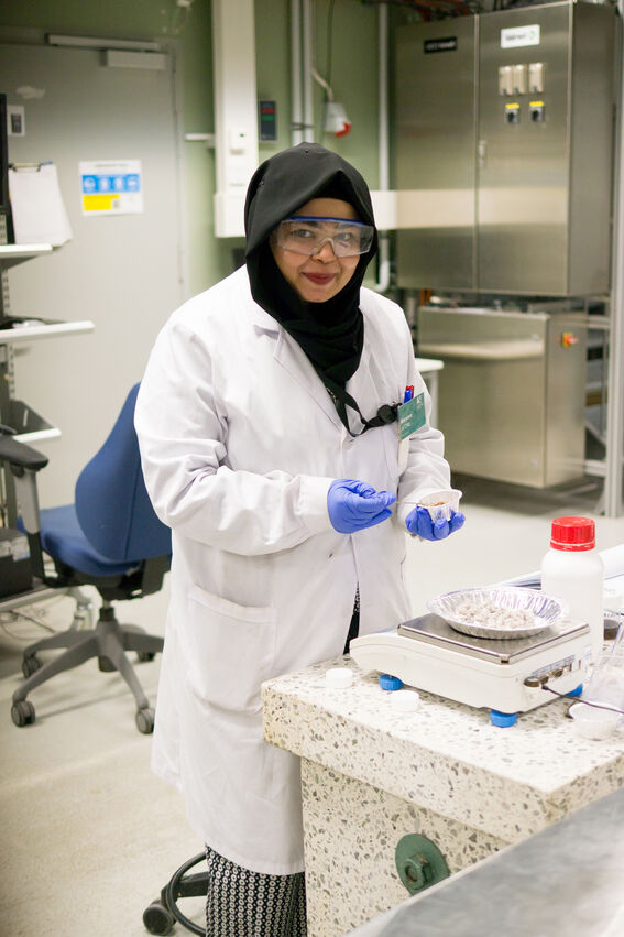 Female scientist in lab coat and hijab weighing samples on a scale in a bright laboratory.
