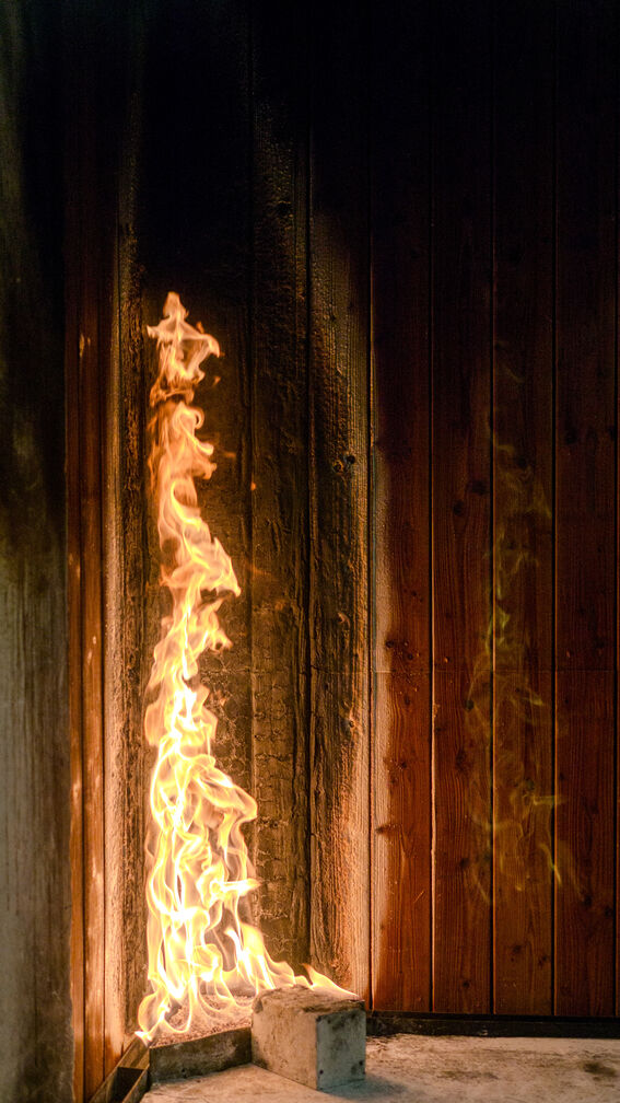 Tall yellow flames climb a dark wooden wall corner beside a concrete block on the floor.