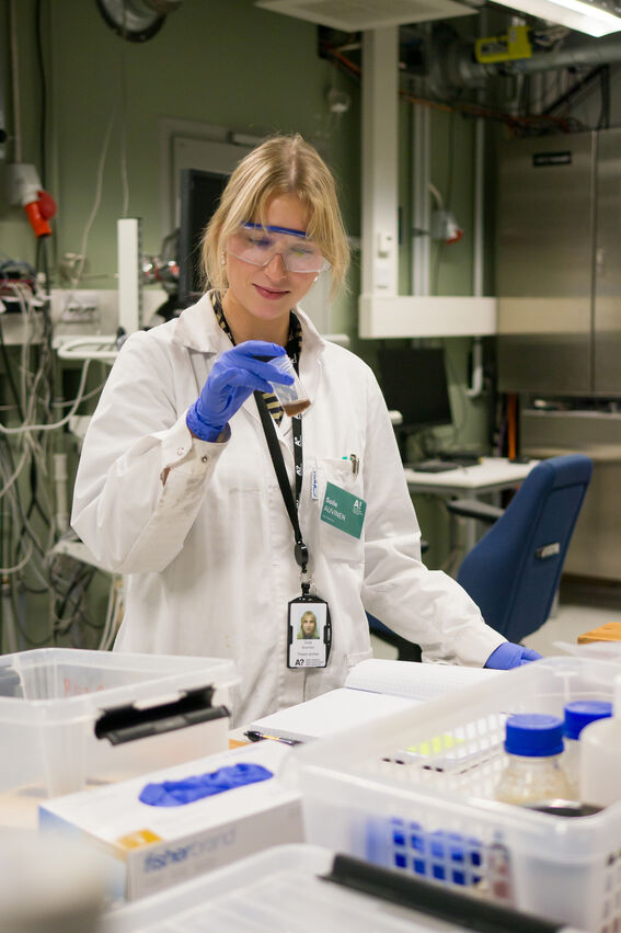Scientist in white coat and blue gloves examining a small sample vial at a busy laboratory bench.