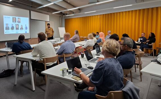 Adult students in a classroom watch a presenter by a screen, using laptops at white tables, yellow curtain behind.
