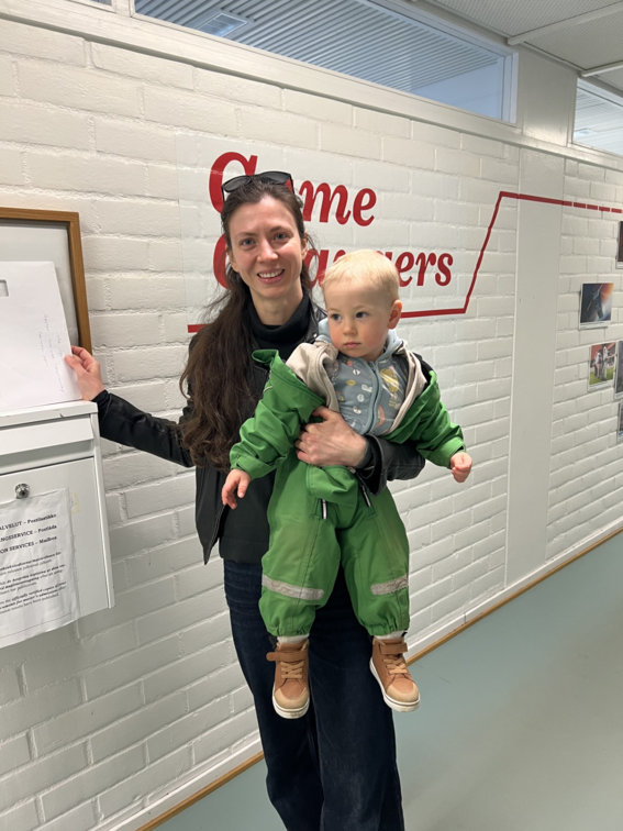 Smiling woman indoors holding a toddler in a green suit, standing by a white brick wall with red text.