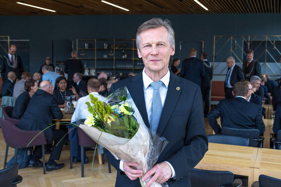Smiling older man in a dark suit holding a flower bouquet at a formal indoor gathering with seated guests.