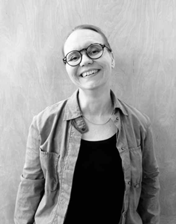 Smiling adult with round glasses in a light shirt over dark top, standing against a plain wall