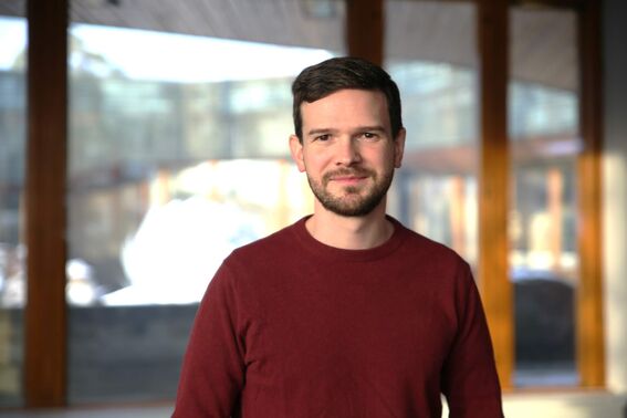 Smiling man with short dark hair in a red jumper, standing indoors before large bright windows
