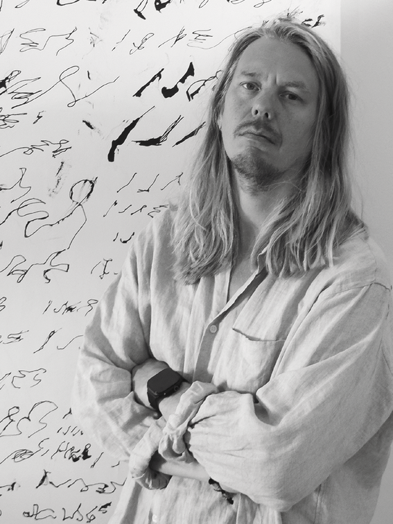 Man in light shirt stands arms crossed before wall of abstract black scribbles.