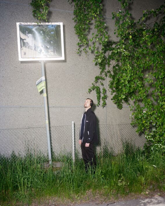 Man in black with headphones stands in tall grass, looking up at a dirty sign by a grey wall and leafy tree