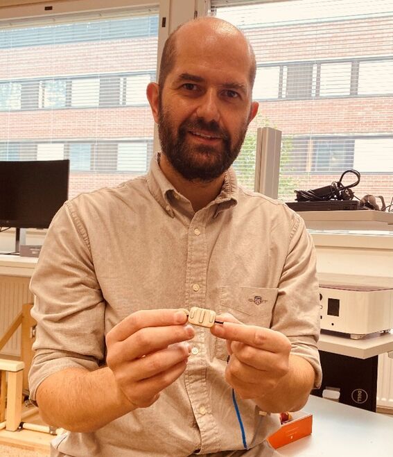 Bearded man in beige shirt sits in office, holding a small beige device with a blue cable