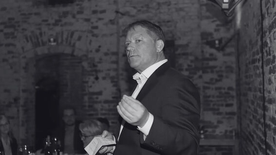 Man in dark suit speaking to diners in a brick-walled room, holding papers in one hand