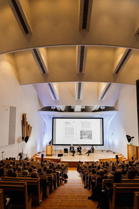 Big lecture hall with wood seats, large screen and panel discussion facing a full audience.