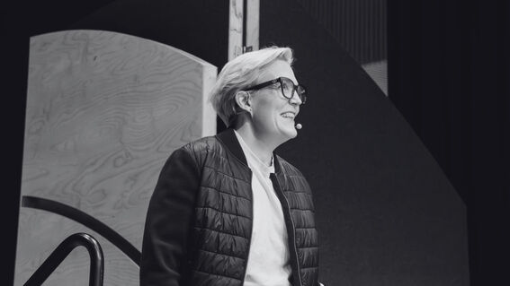 Smiling person with short light hair and glasses speaking on stage in a dark jacket, monochrome photo