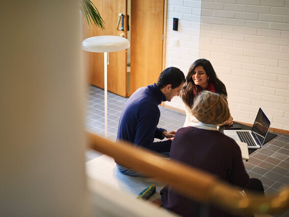 Kolme ihmistä istuu lattialla käytävässä kannettavan kanssa keskustellen.