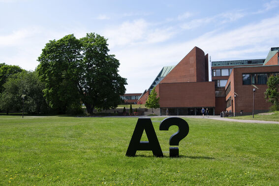 Aalto University logo with question mark on grass, with a campus building visible in the background