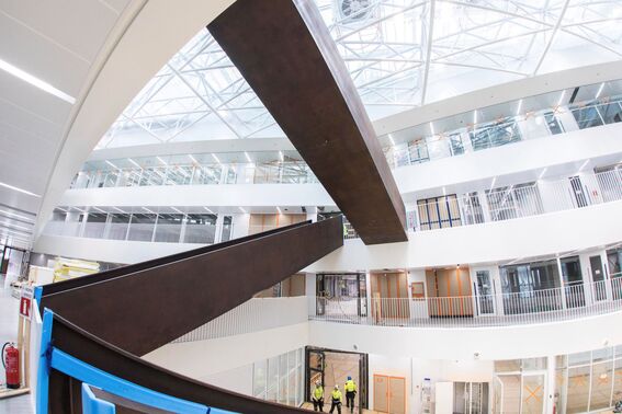 Moderni atrium toimistorakennuksessa, jossa on kaksi tummaa metallipalkkia, suuret ikkunat ja ihmiset turvavarusteissa.