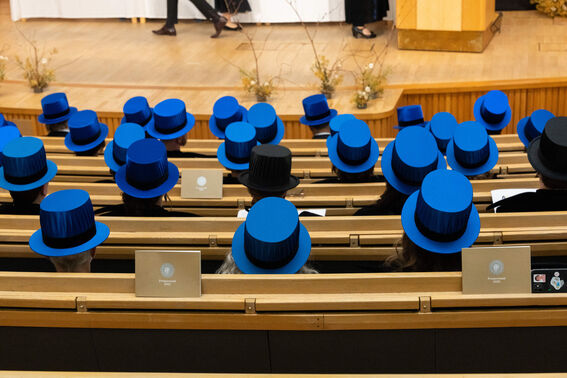 People sitting in an auditorium wearing bright blue top hats, with two individuals in black hats, facing a stage.