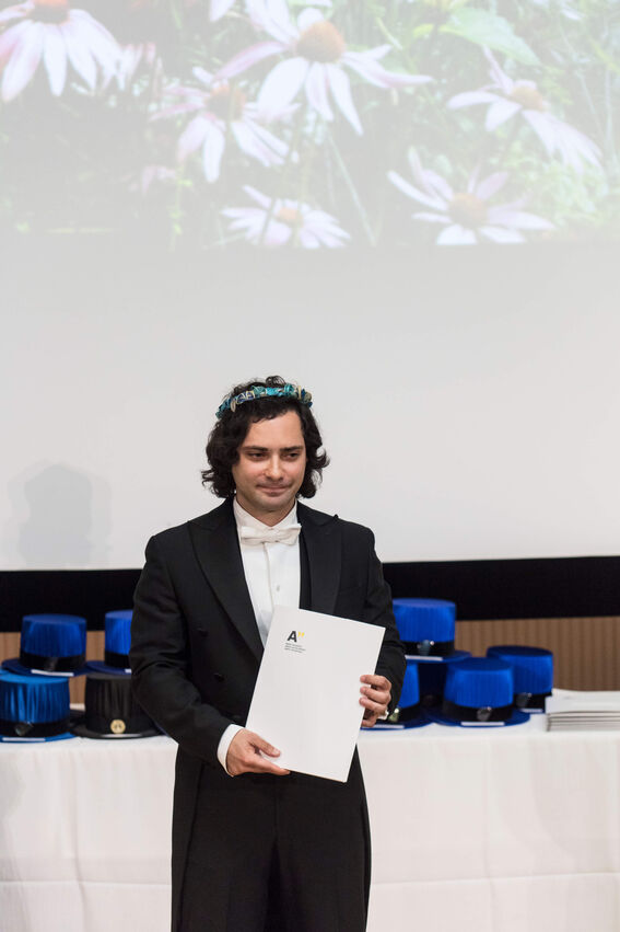 A person in a black suit holds a certificate with the letter 'A'. Background shows a stack of blue hats and a flower projection.