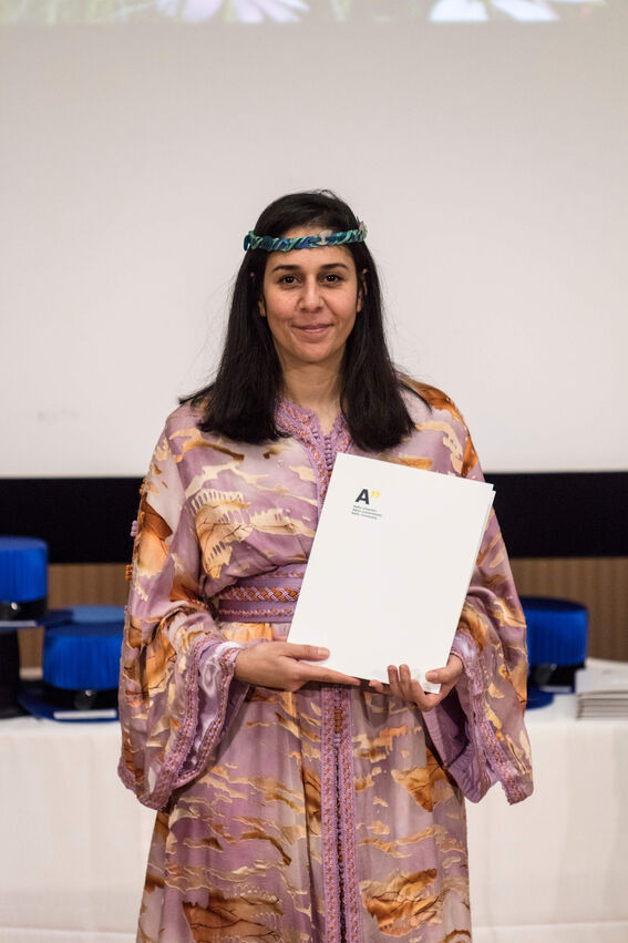 A person with black hair holding a certificate. They wear a purple floral dress with long sleeves.