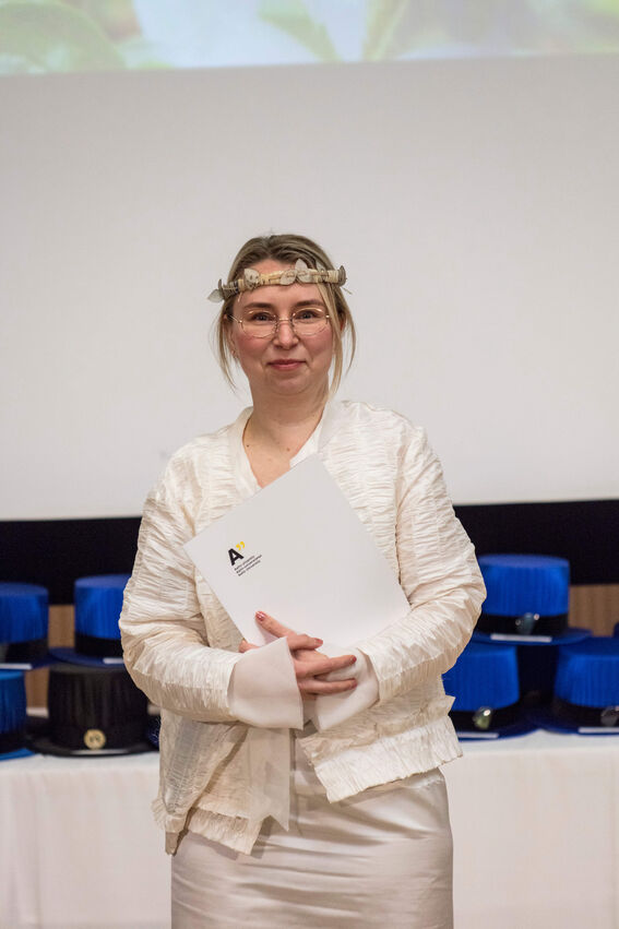 Person wearing a white outfit holding a certificate from Aalto University in front of blue doctoral hats