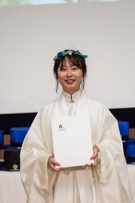 Person wearing a white robe and blue flower crown, holding an Aalto University certificate.