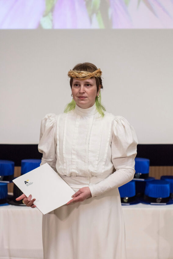 Person in white dress with puffed sleeves, holding a certificate. Blue caps in the background.