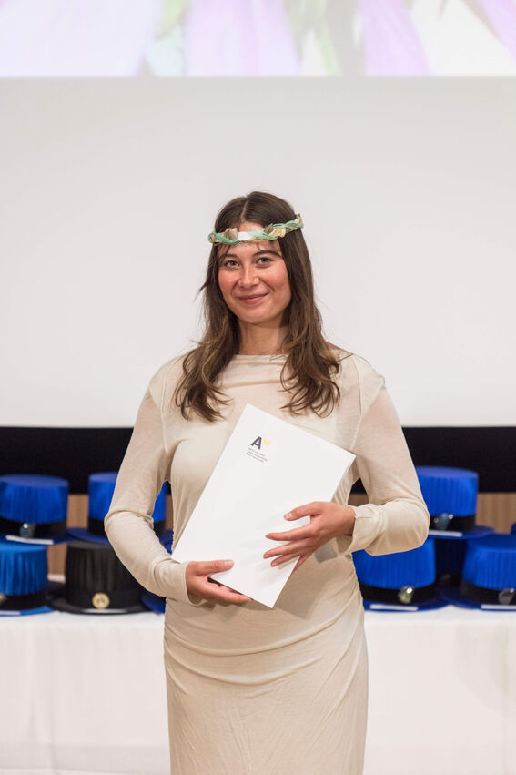 A woman in a light dress holds a diploma in front of a display of doctoral hats with blue tassels.