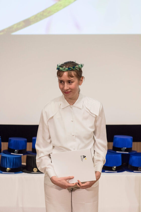 Person dressed in white holding a certificate, standing in front of a table with blue and black hats.