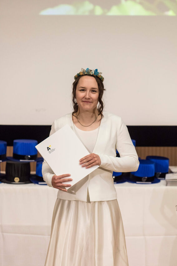 A person holds a folder while wearing a white dress and jacket. They have a colorful wreath.