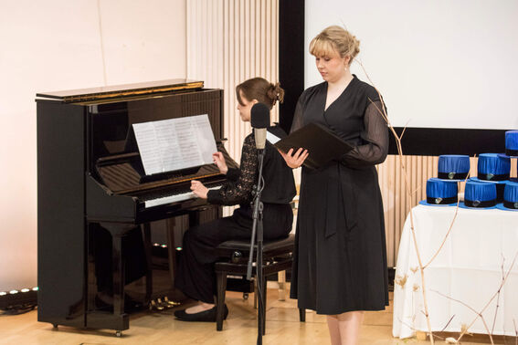 A person plays the piano while another stands holding a book. Several cylindrical blue awards are on a table next to them.