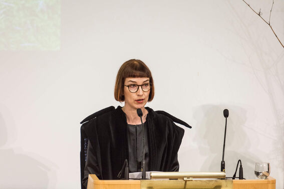 Person wearing a dark robe, standing at a podium with two microphones and a glass of water.