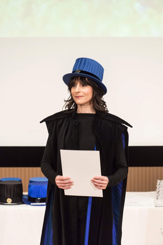 Person in academic robes and a blue doctoral hat holding a white diploma.
