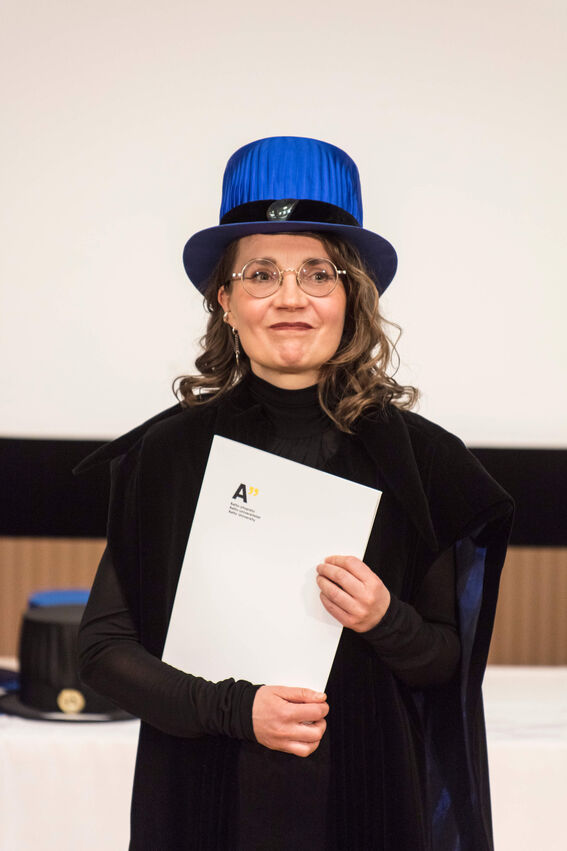 Person wearing a blue doctoral hat and black clothes, holding a diploma from Aalto University.