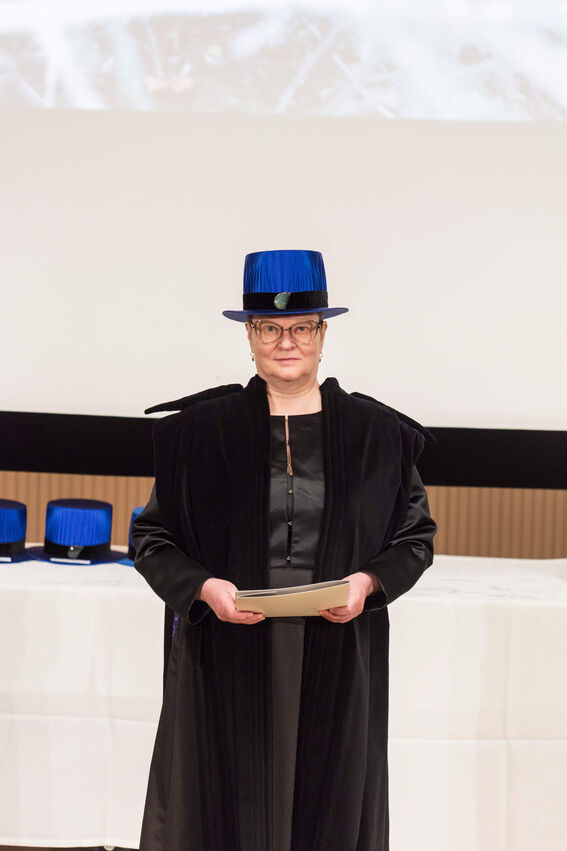Person wearing a black academic gown and a blue academic hat, holding a certificate with more blue hats in the background.