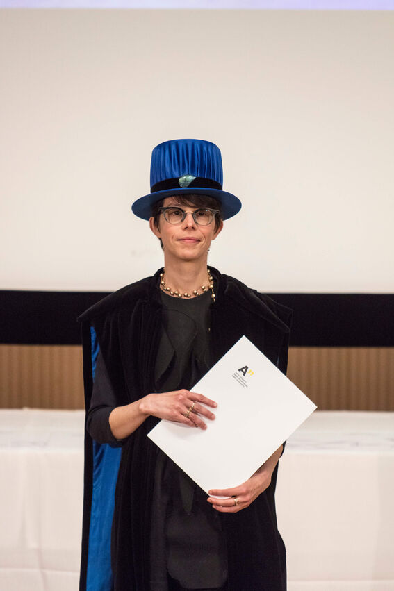 Person wearing ceremonial attire with a blue hat, holding a diploma in front of a screen