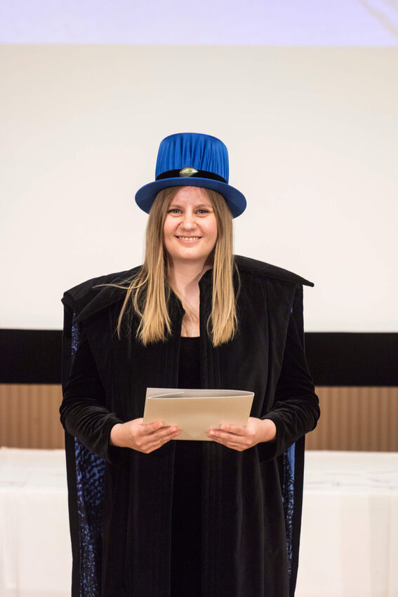 Person wearing a blue hat and black robe, holding a document indoors with a presentation screen background.