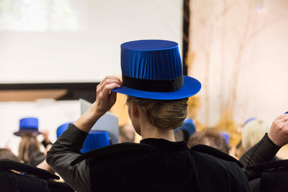 A person in formal attire adjusts their blue ceremonial hat in an auditorium filled with others wearing similar hats.