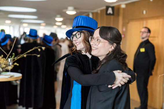 Two women hugging at an academic ceremony, one wearing a blue hat and cape.