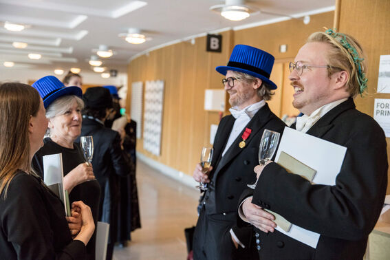 Several people standing indoors, some wearing academic regalia with blue hats, holding glasses and documents.