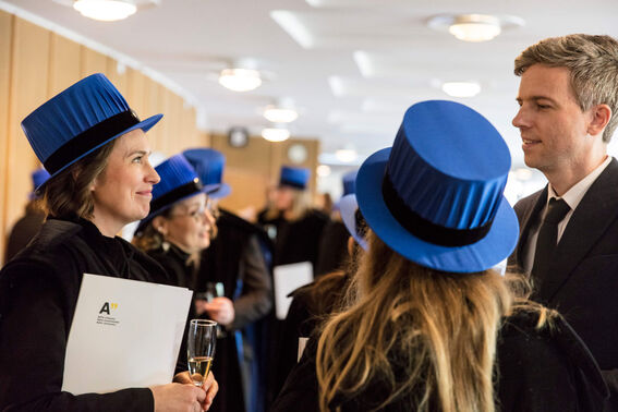 People wearing blue hats and black gowns at an academic event, holding diplomas and glasses.