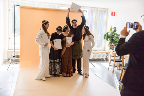 Five people in traditional attire holding certificates, posing indoors with an orange backdrop while one person takes a photo.