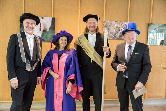 Four people dressed in ceremonial academic attire, standing indoors with artwork on the walls.