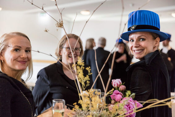 Three people in dark attire converse near a floral arrangement with pink roses and dry branches. One wears a blue hat.