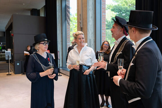 A group of people in formal attire holding glasses of champagne indoors. Some wear academic ceremonial attire.